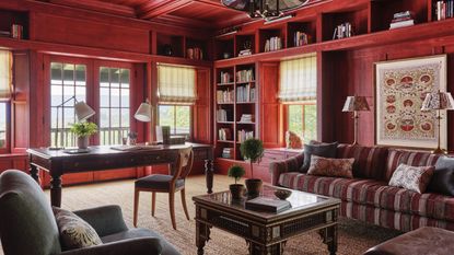a vast home library clad in wood and stained red with a large antique desk, upholstered patterned couch, a tapestry on the wall and layered lighting