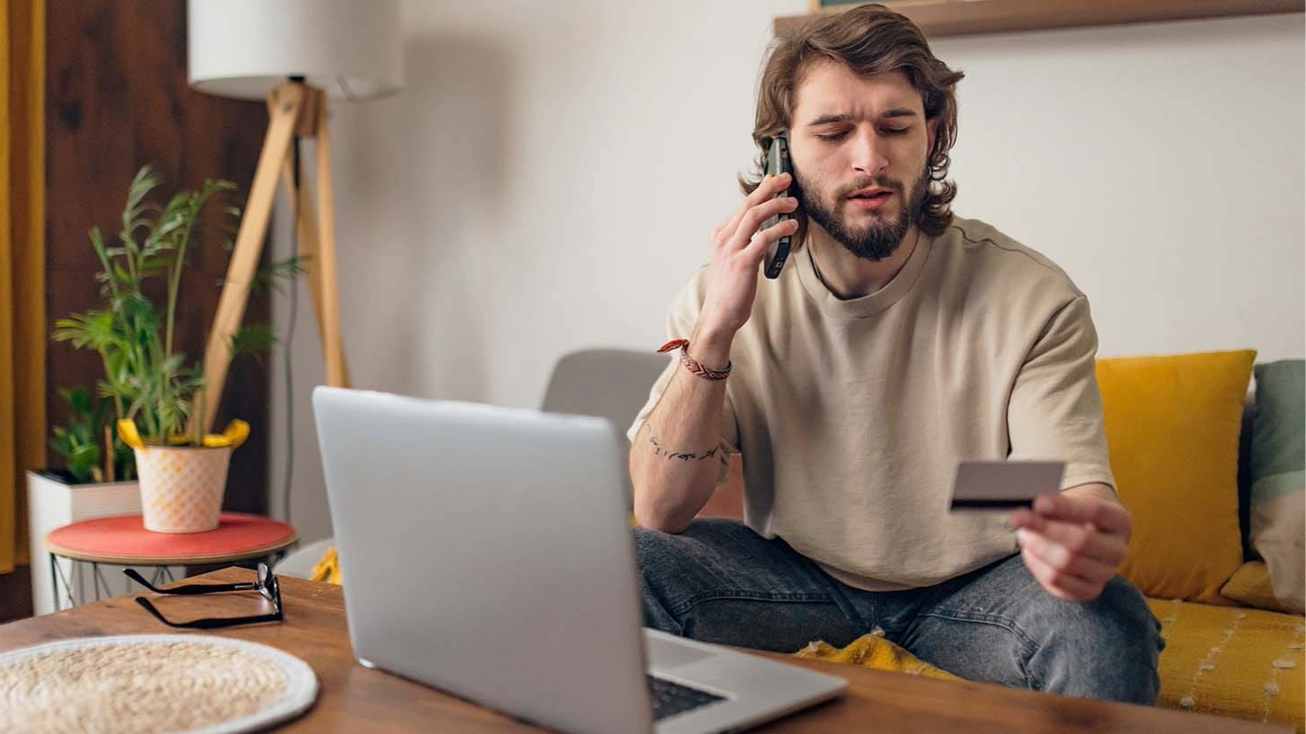 A man looking at his credit card. He's on the phone with his laptop open in front of him.