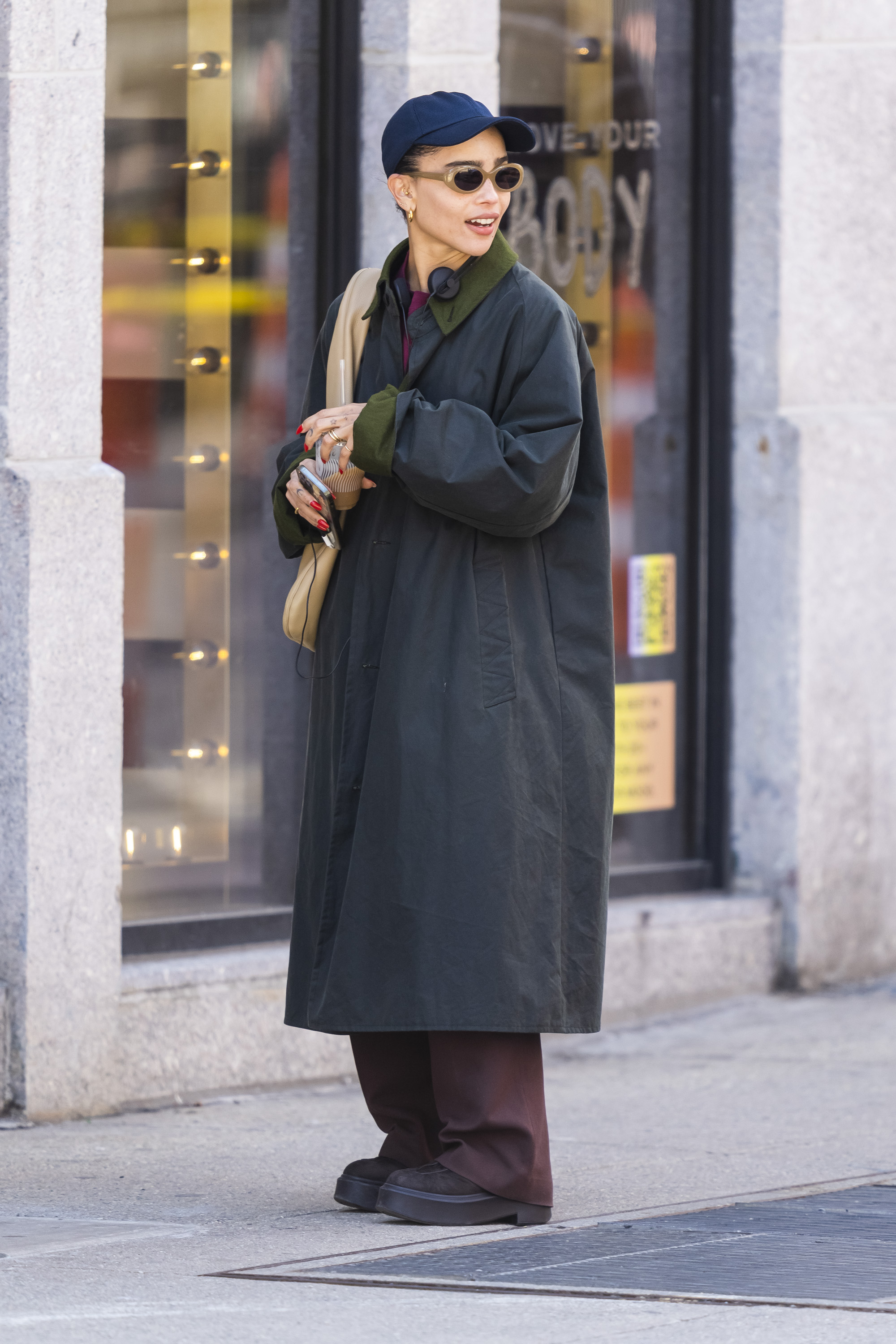 Zoe Kravitz wears a long black coat, brown pants, brown boots, and a navy baseball cap.