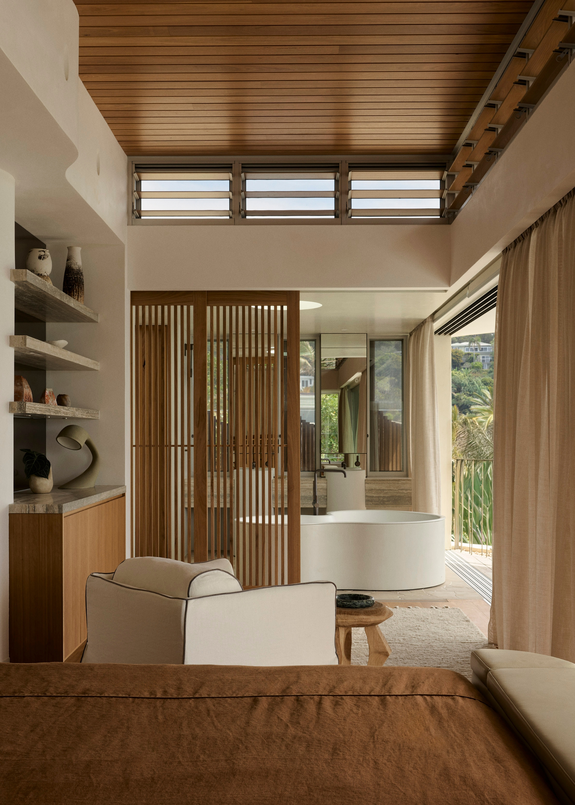 An earthy bedroom with a white armchair, a wood side table, a deep rust bedscape, and a slatted door leading to a bathroom with a freestanding tub