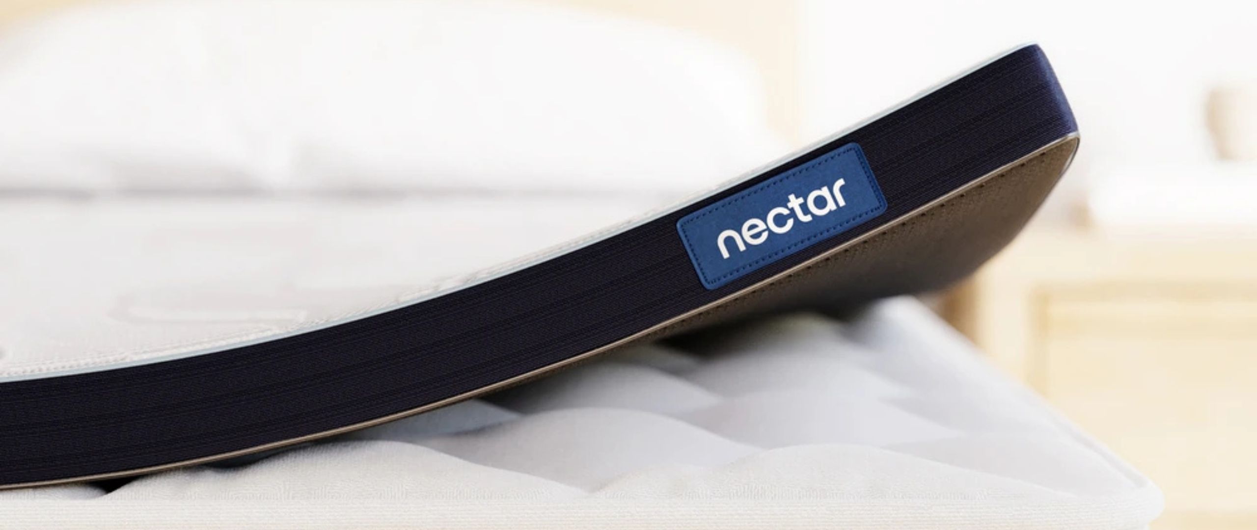 The foot of the Nectar Mattress Topper, with the &quot;nectar&quot; logo, slightly curled at the bottom right-hand corner and placed on top of a white mattress. 