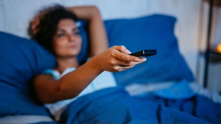 Woman laying in smart bed with TV and blue bedding propped up by pillows reaching out with a remote control.