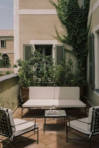 A hotel room features a lush terrace filled with plush, wrought-iron sofas and couches and traditional Mediterranean style facades.