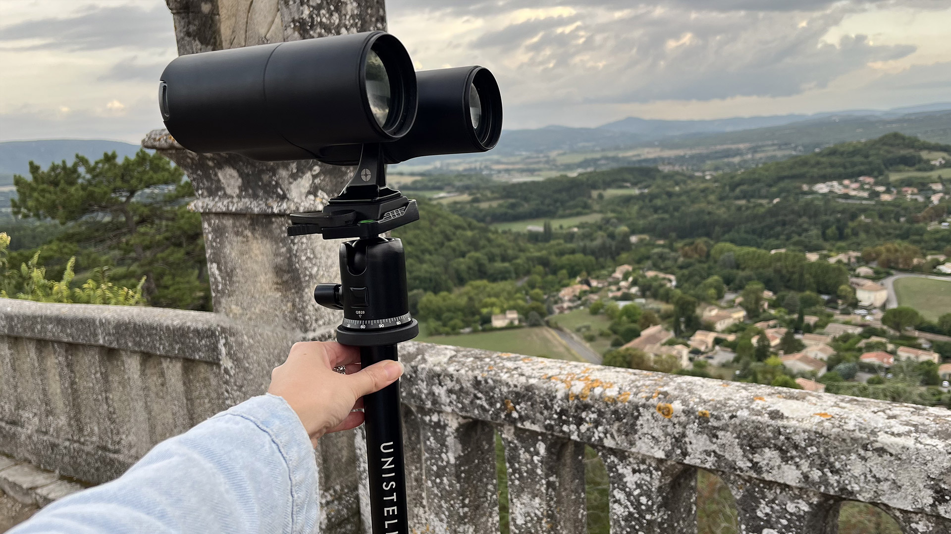 A hand holds a monopod that has a pair of binoculars mounted, with a country vista in the background