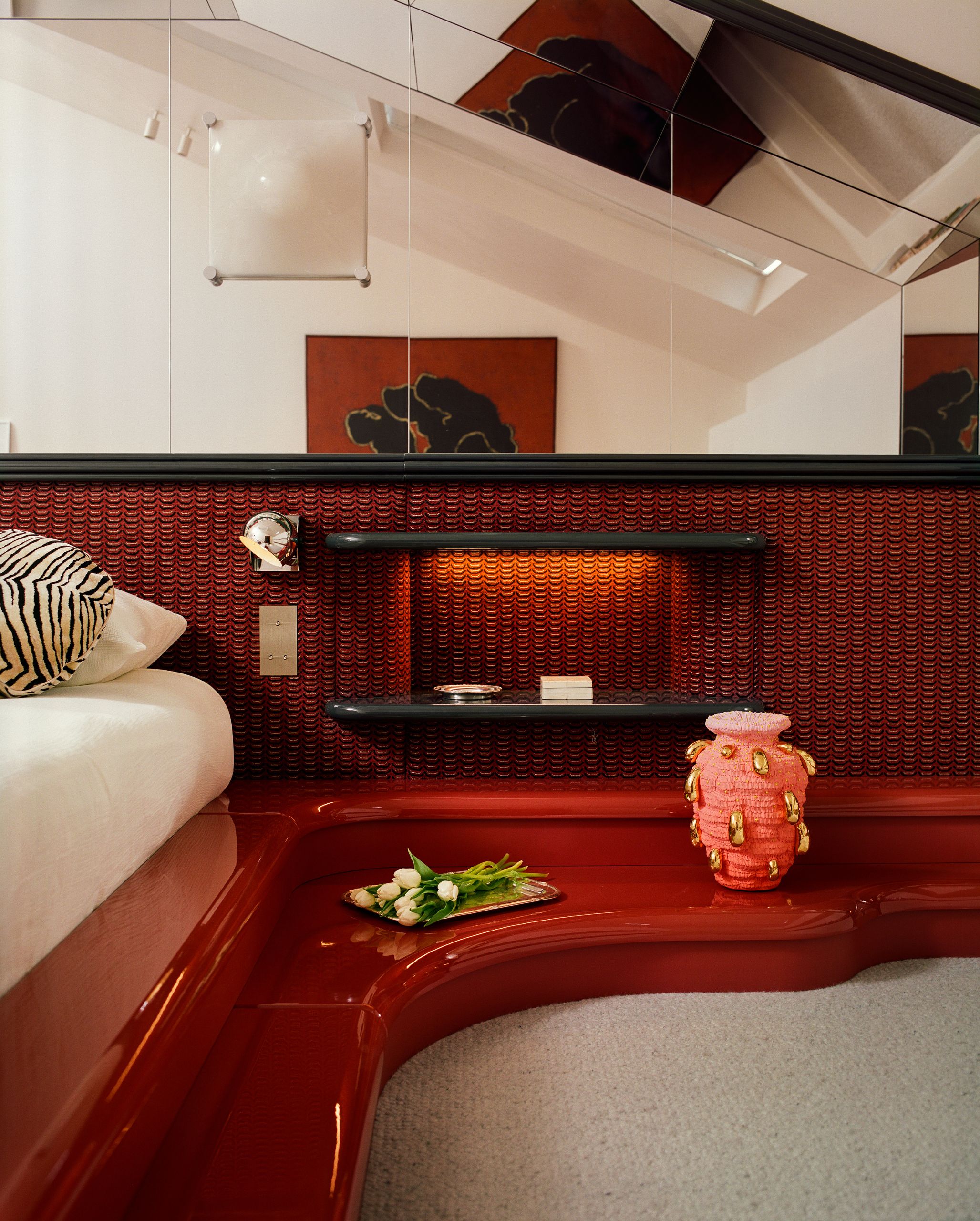 A bedroom with a built-in bed that has red lacquer steps up to it that wrap around the wall. The wall is a red fabric with mirror on the upper half. There is a zebra pillow on the bed.