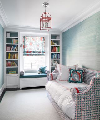 Blue kids playroom with alcove white book shelves, bench seat beneath window with patterned colorful blind, blue and red patterned upholstered daybed with cushions and duvet