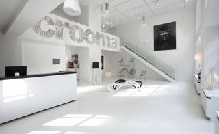 Interior room, white floor ceiling, walls, floor, furniture and accessories with black trim, large Crooma sign, black and white portrait image on right wall, computer screen, stacked chairs, lighting