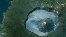 A satellite photo showing a volcano inside a massive crater lake with clouds reflected perfectly in the water's surface