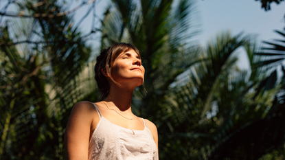 Woman with closed eyes and sunshine on her face with palm trees behind her - she looks like she could have used one of the best fake tans