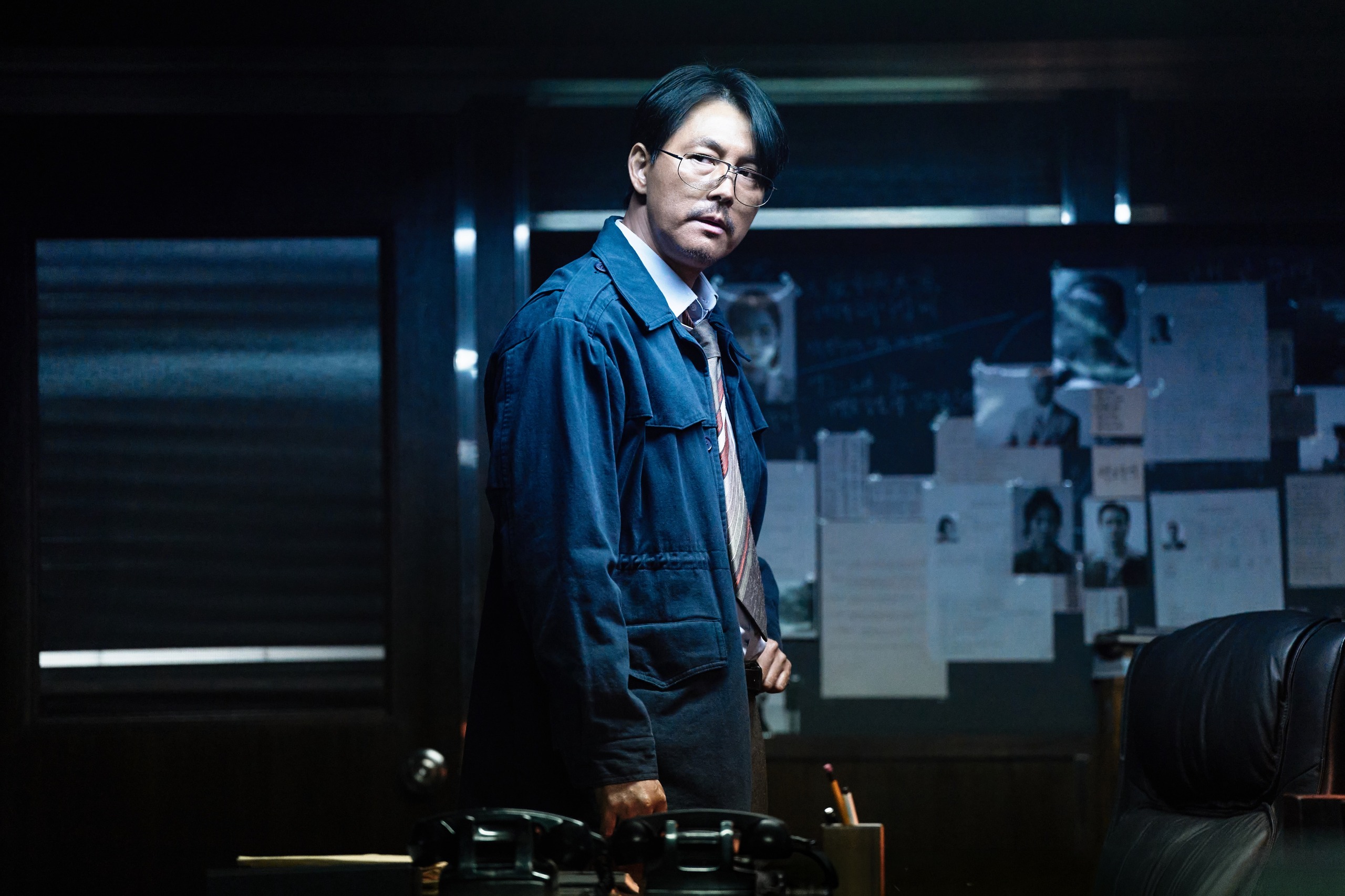 A Korean man in glasses and a blue jacket (Jung Woo-sung as Jang Geunyoung) holds a briefcase while standing in a dark office in front of an investigation board, in Hulu's 'Made in Korea' season 2.