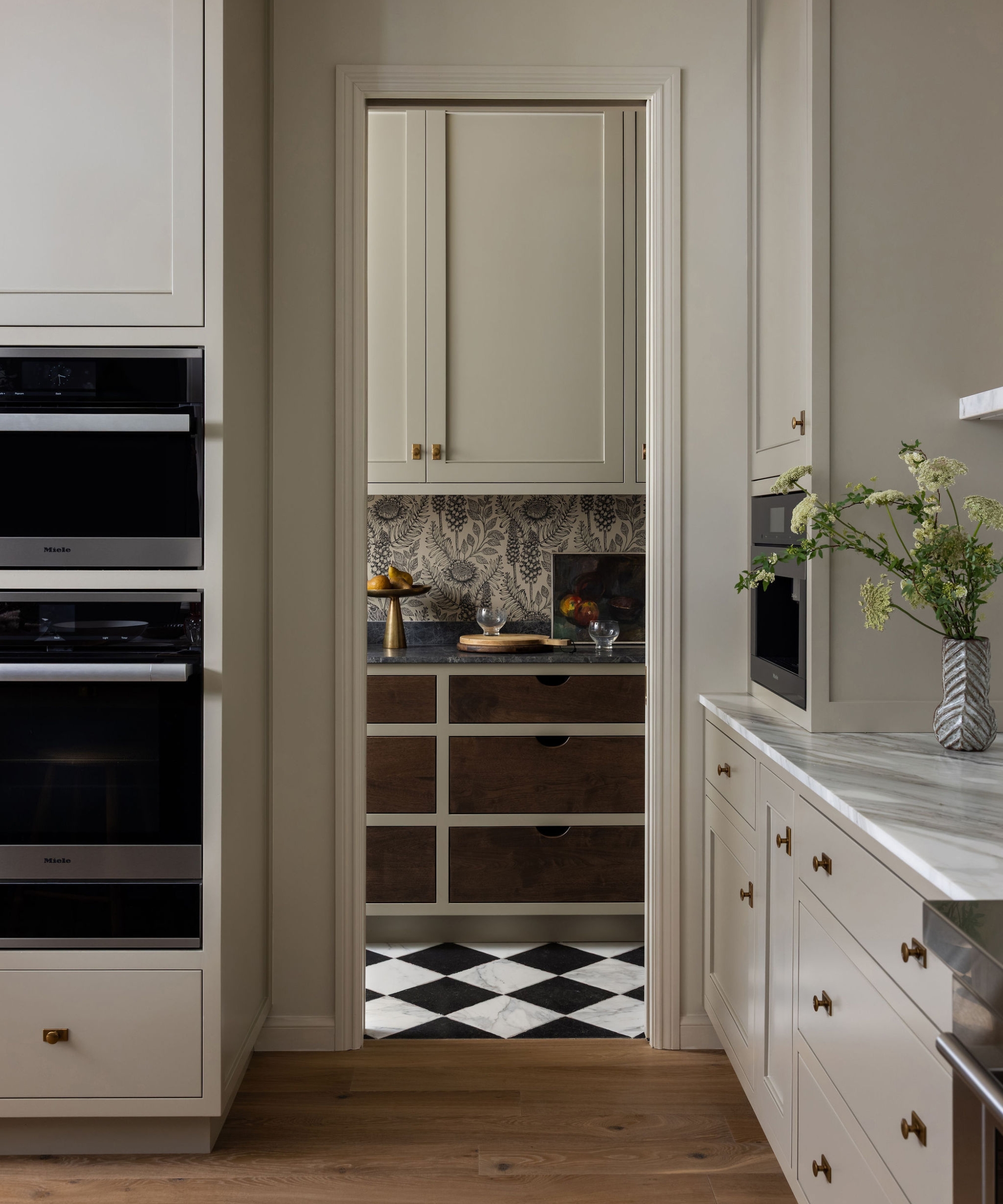 Pantry viewed through the kitchen doorway, with botanical wallpaper, wood drawers, pale cabinets and a black-and-white floor.