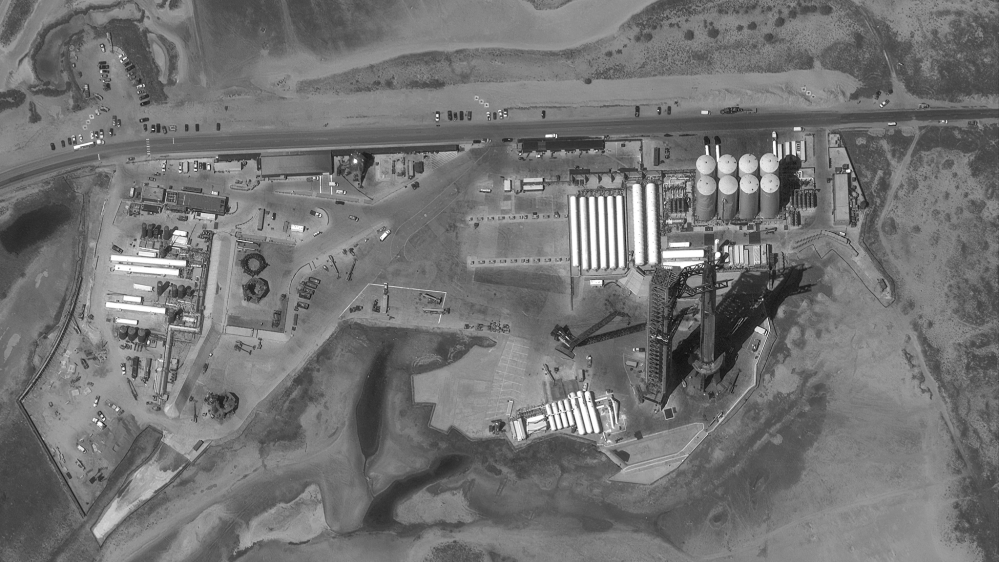 black-and-white satellite photo of a large rocket standing next to a launch tower with dirt and white buildings in the background
