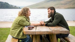 Helen (Danielle Macdonald) and Elliot (Jamie Dornan) hold hands across a picnic bench in front of a lake in The Tourist season 2
