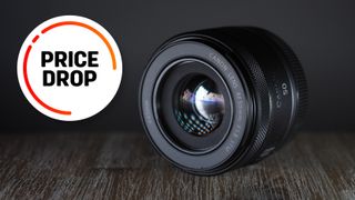 Canon RF 50mm f/1.8 lens on a wooden surface, in front of a dark shadowy background, with the text "Price drop"