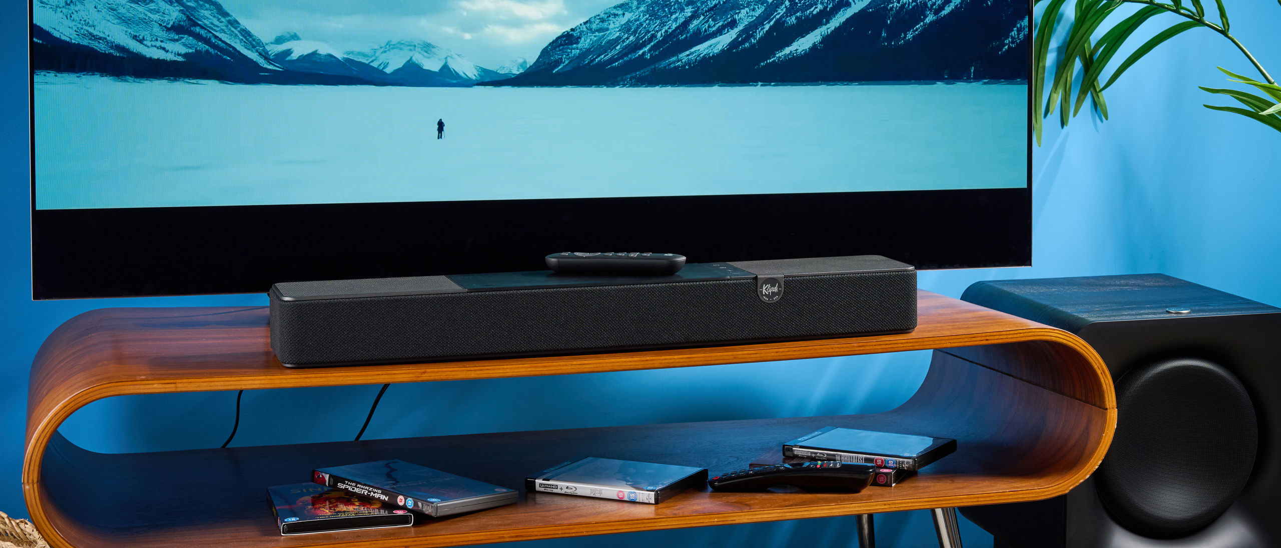 the klipsch flexus core 100 soundbar photographed on a wooden tv stand with a tv behind, showing the remote, buttons, and inputs