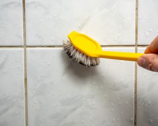 Person with brush and yellow glove cleaning grout on a shower wall