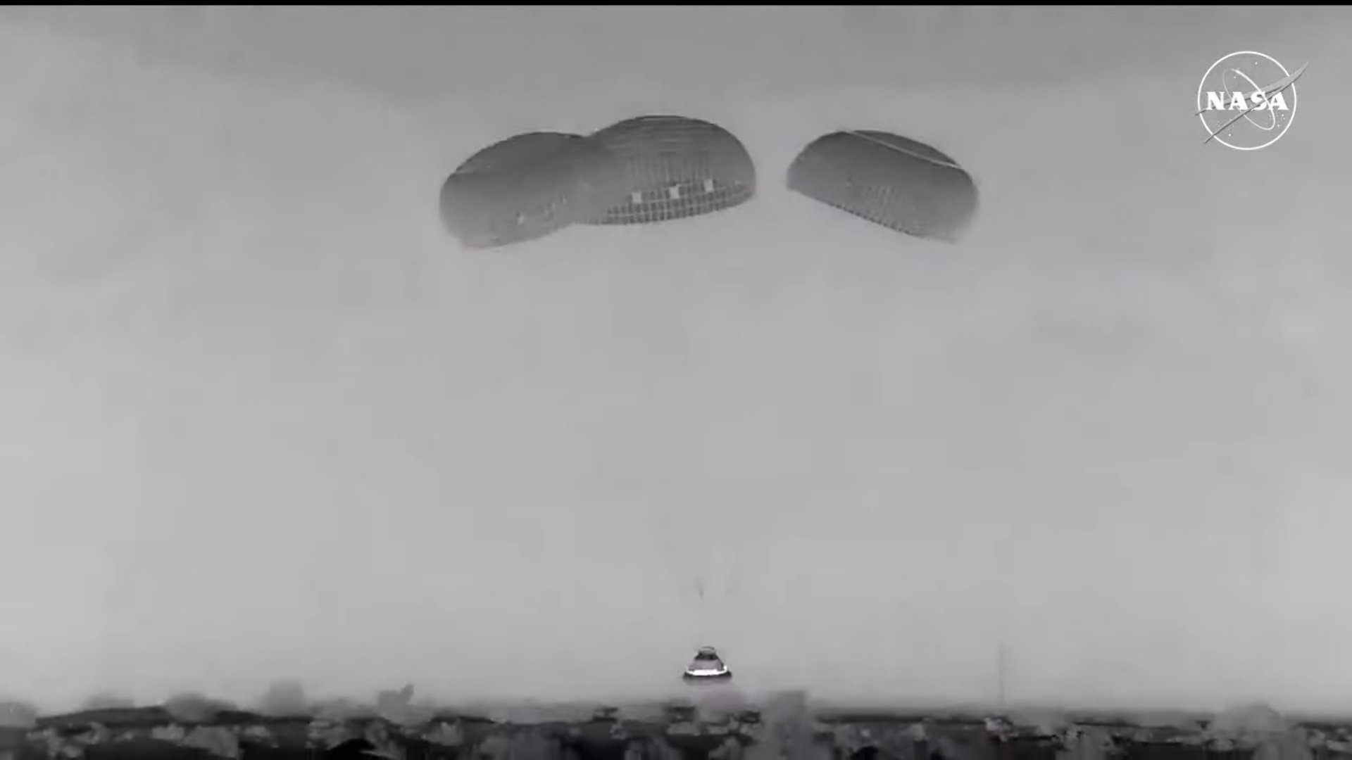 A black and white view of a capsule descending under parachutes. A NASA logo can be seen at the top right as the show was on NASA television