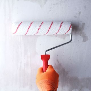 Person in an orange glove using a roller to mist coat white wall