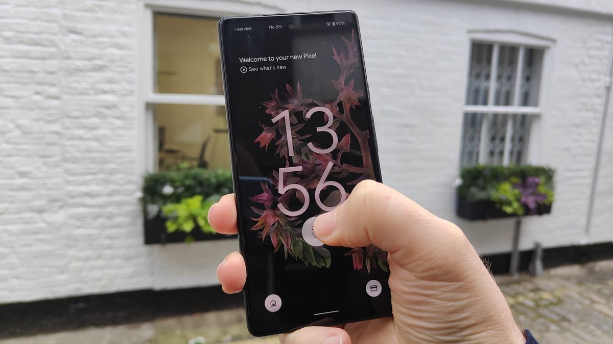 The Google Pixel 6 in Stormy Black, with a thumb on its fingerprint sensor, in front of a white brick background