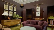 living room with chocolate walls, green rug, purple sofa, light blue fringed ottoman, blush pink armchair, timber console with lamps, and artworks hanging from a brass rail