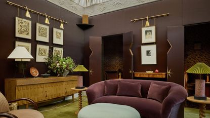 living room with chocolate walls, green rug, purple sofa, light blue fringed ottoman, blush pink armchair, timber console with lamps, and artworks hanging from a brass rail