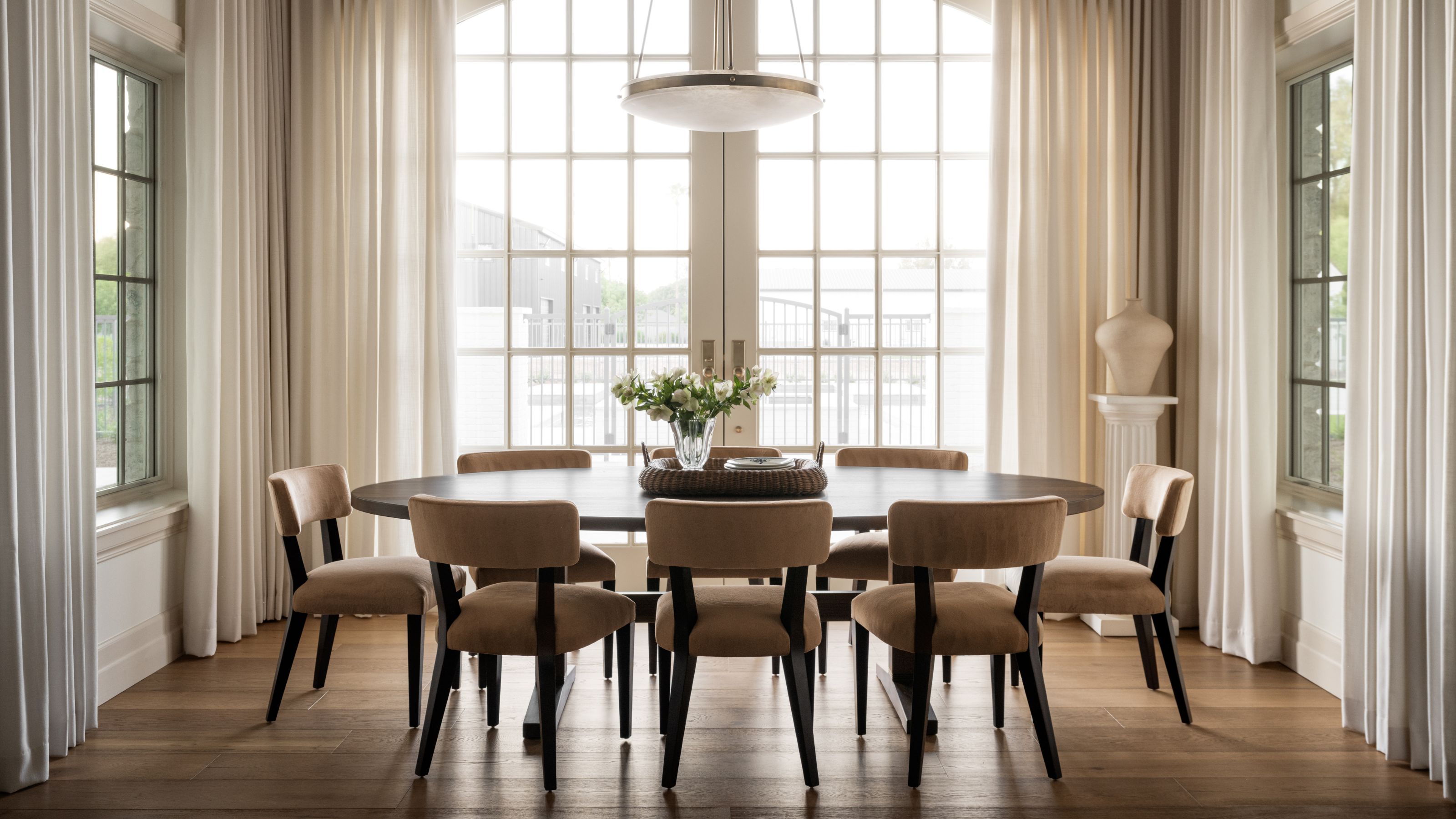 a dining nook in an opening plan living kitchen area with large glass doors and windows framed by drapes and a simple oak table and black upholstered chairs