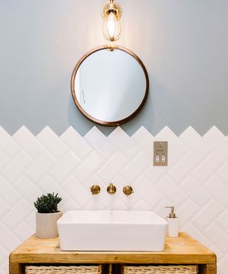 An example of bathroom lighting trends showing a white sink below a round mirror and wall light