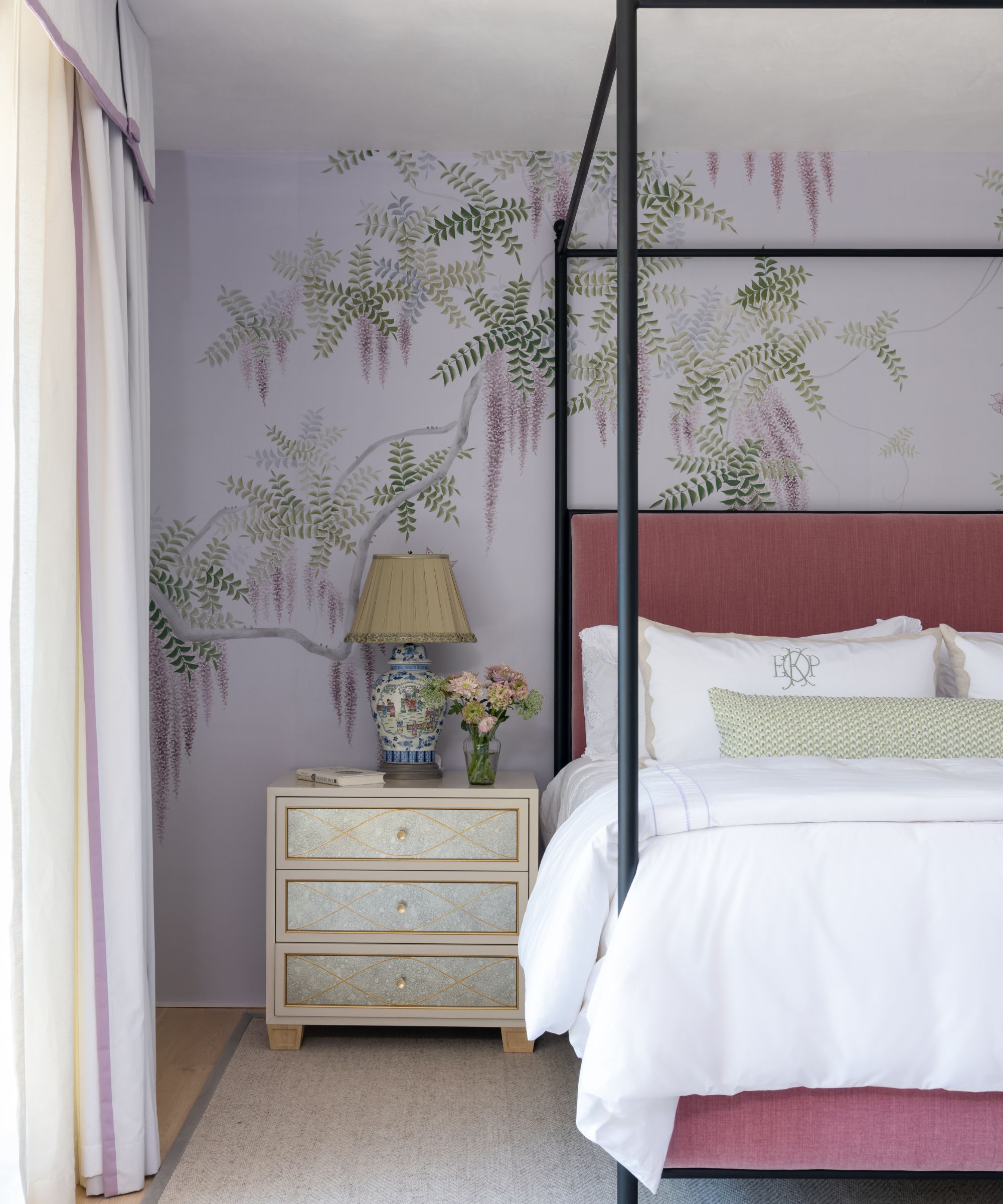 A bedroom with a lilac floral wall mural, a four-poster bed with a mauve headboard, and crisp white bedding. Neutral carpeted floor and a neutral nightstand with an antique lamp.