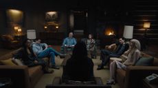 Production still from Netflix's show 'Something Very Bad is Going to Happen'. There are seven people sitting in a dark, mid-century-style living room on multiple sofa with lots of lamps around the room.