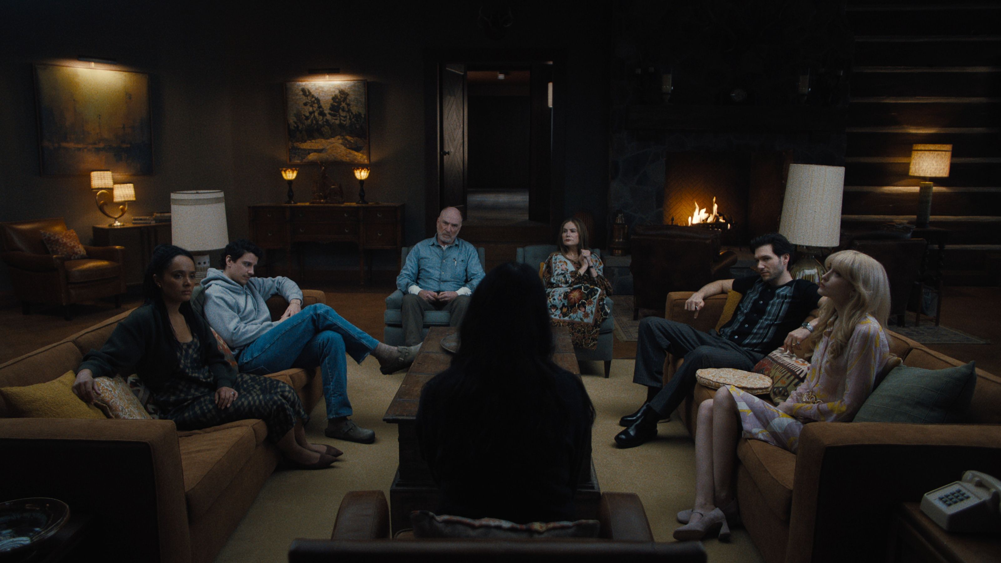 Production still from Netflix's show 'Something Very Bad is Going to Happen'. There are seven people sitting in a dark, mid-century-style living room on multiple sofa with lots of lamps around the room.
