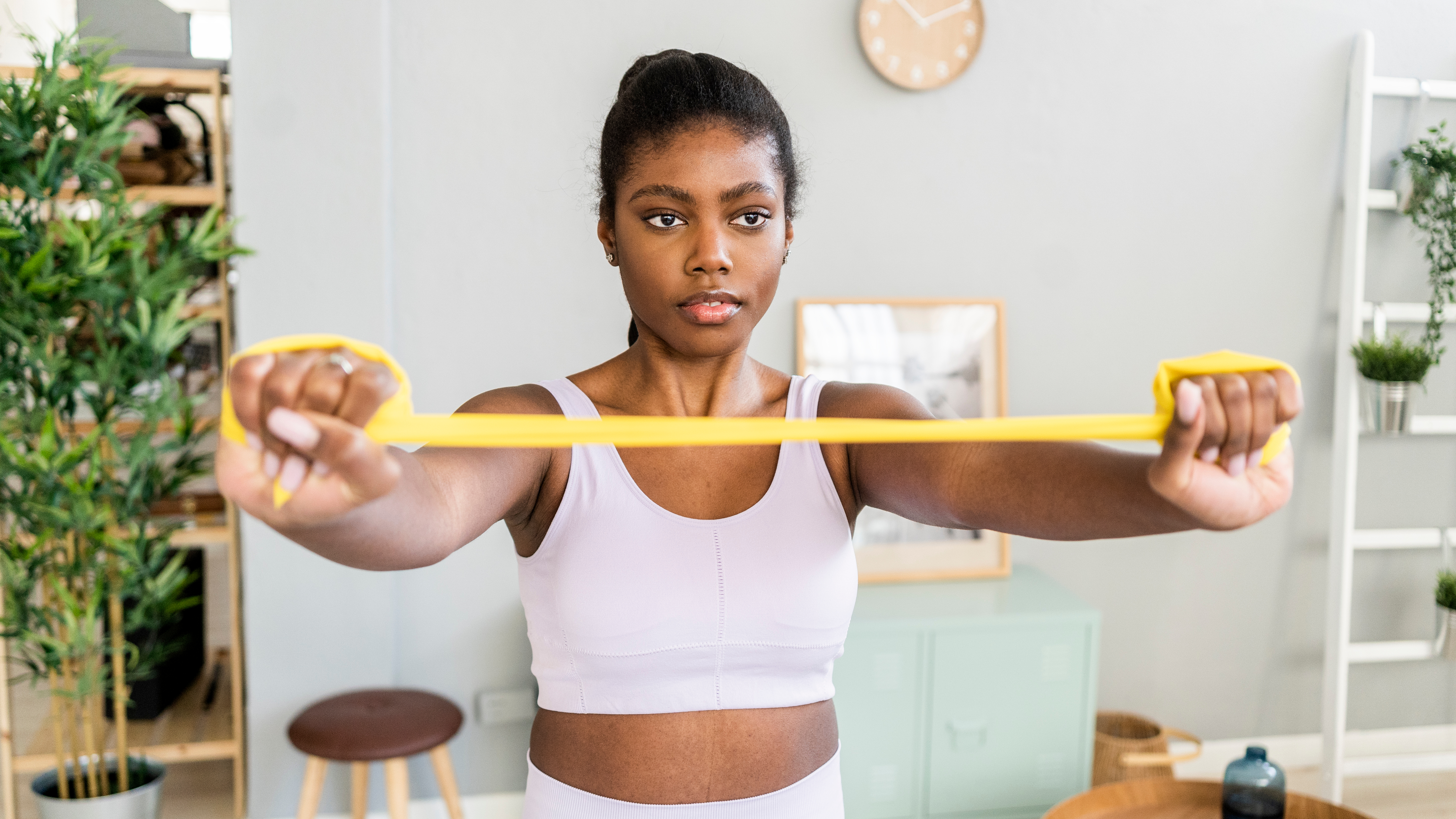 A woman in a sports bra and leggings stands in a living room holding a resistance band taut between her hands, which are outstretched in front of her. Behind her we see a stool, shelving and plants.