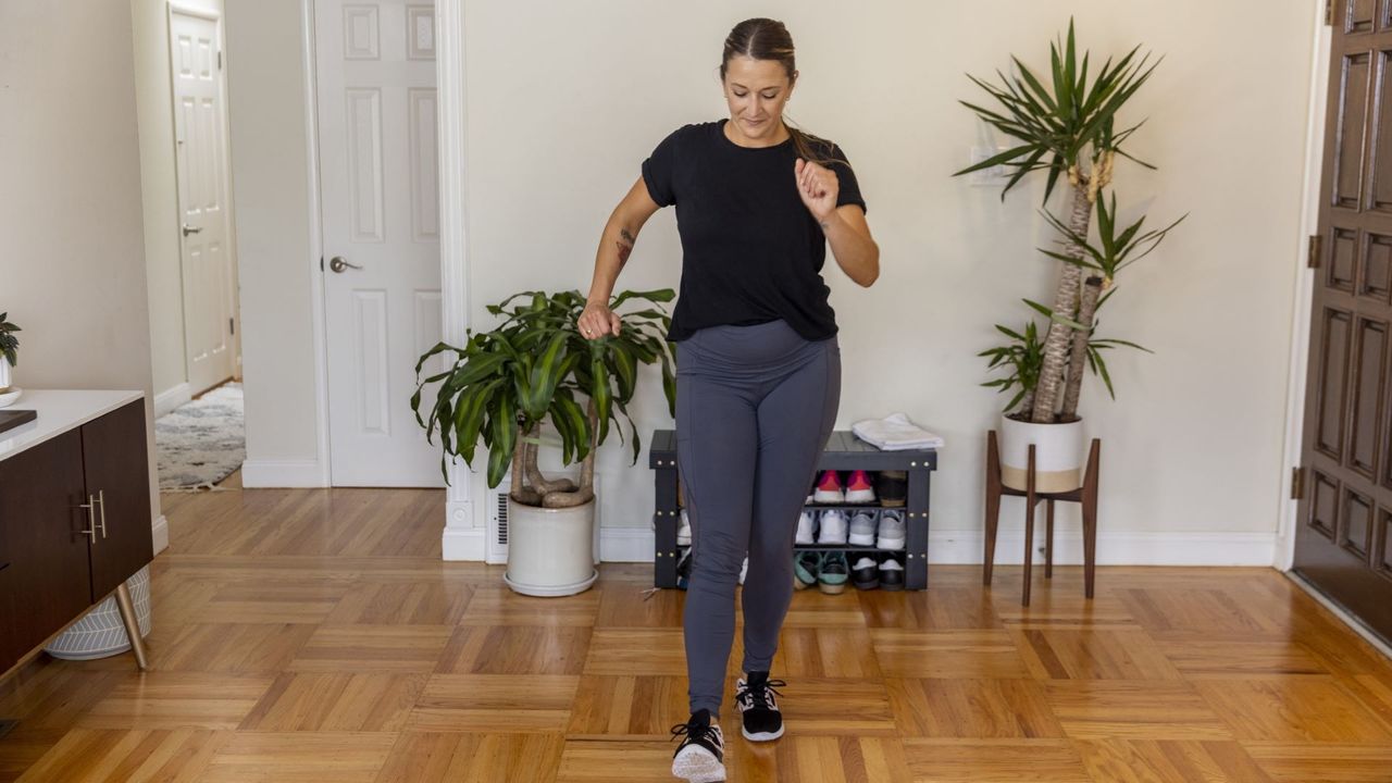 A woman in grey leggings and a black t-shirt does a walking workout in her living room.