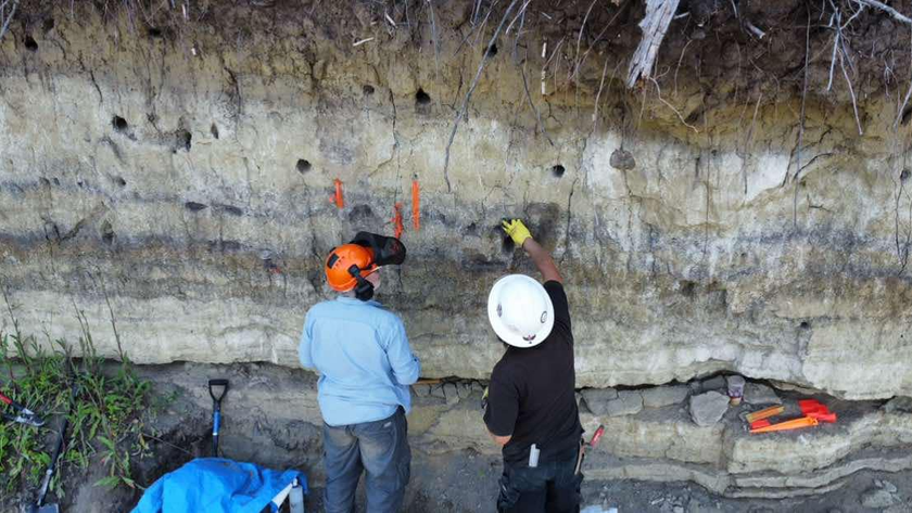 Two people standing in front of a cliff, looking at the different colored lines of soil running horizontally