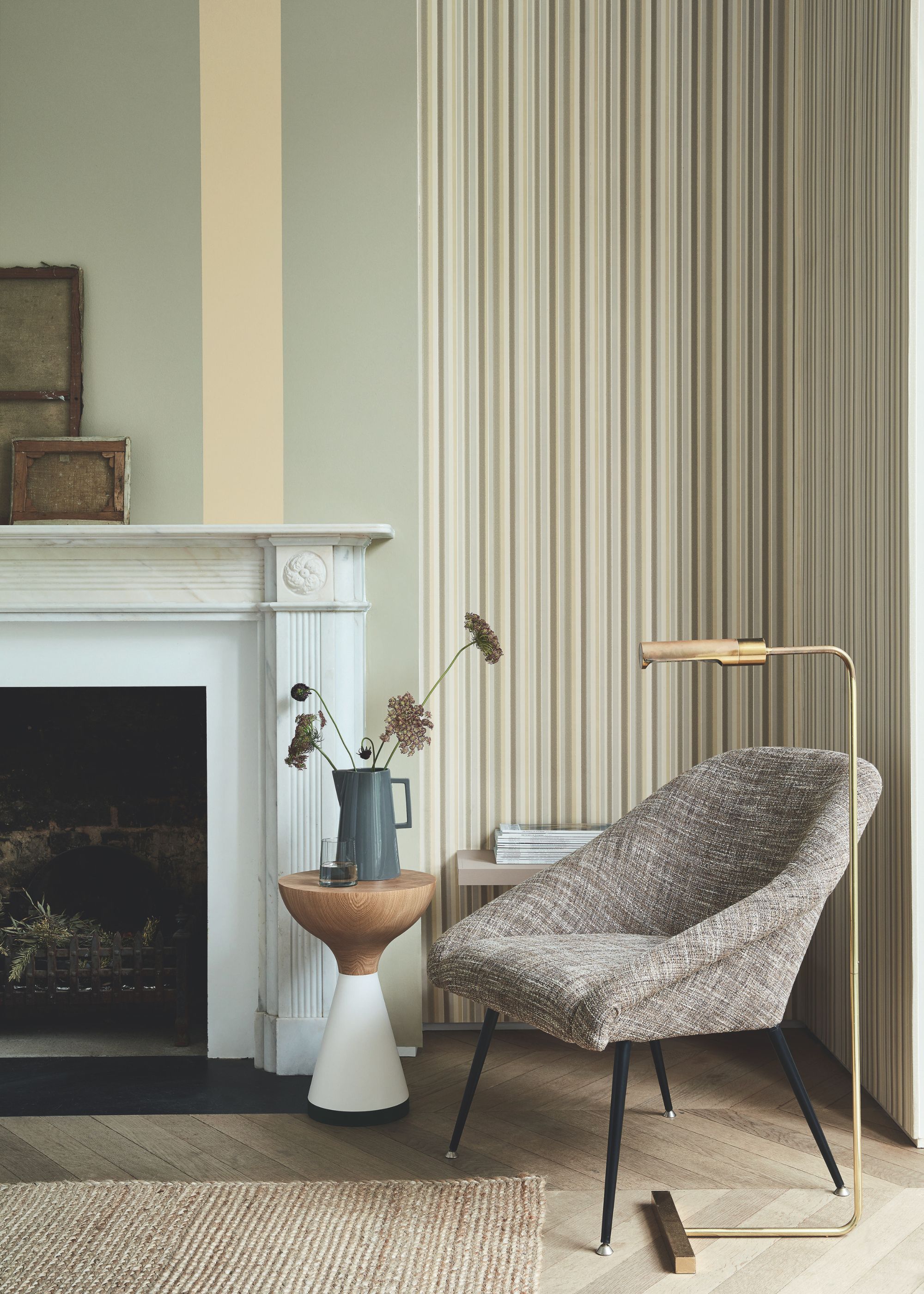 A traditional living room with soft light green walls with striped wallpaper in the alcove. A white fireplace, parquet flooring with a jute rug, and a gray modern armchair with a brass lamp.