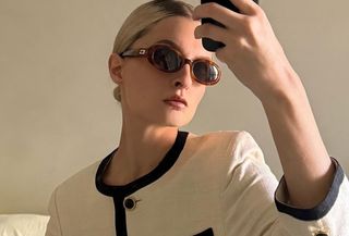 Woman wears white and black jacket, and brown sunglasses while talking mirror selfie.