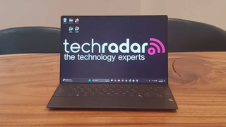 The Dell XPS 13 on a wooden desk in an office during our tests, the screen displays the TechRadar logo as the wallpaper