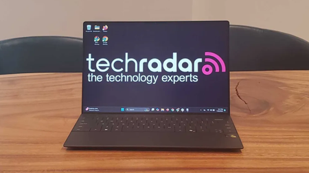 The Dell XPS 13 on a wooden desk in an office during our tests, the screen displays the TechRadar logo as the wallpaper