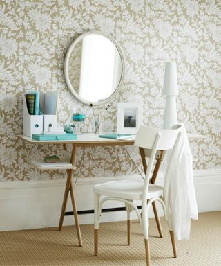 An office area with white and beige flowery wallpaper, a white circular mirror, a wooden desk with blue books, a white photo frame, and white file holders, with a white chair in front with a white shirt on it