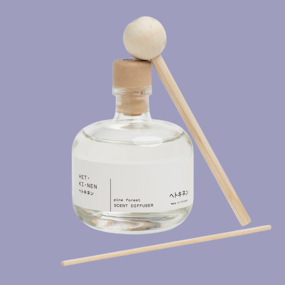 A transparent, chubby bottle of home fragrance with a minimalist white label and organic cork is placed against a lilac background along with two fragrance sticks.