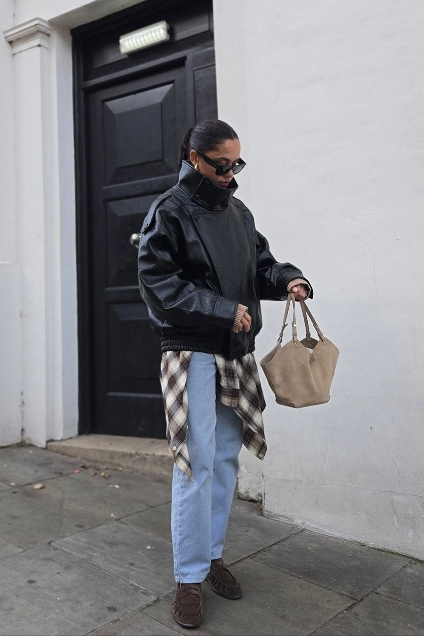 A woman standing outside in front of a black door wearing a black funnel neck leather bomber jacket styled with a tan and white flannel peaking out underneath, light wash straight-leg jeans, dark brown suede loafers, a light tan suede handbag, gold earrings, and black rectangular sunglasses.