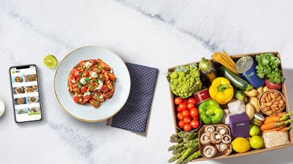 A box full of fresh fruit and vegetables, a bowl filled with a tomato-based pasta dish and a phone showing the HelloFresh app