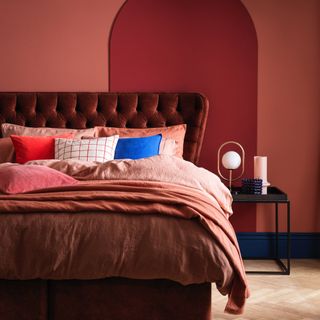 small bedroom colour ideas, red bedroom colour scheme with dark red velvet bed, coral bedding, herringbone floor, black side table, modern lamp, vases, red and blue cushions
