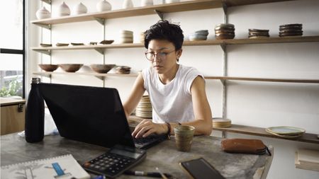 A small business owner works on her laptop in her pottery shop.