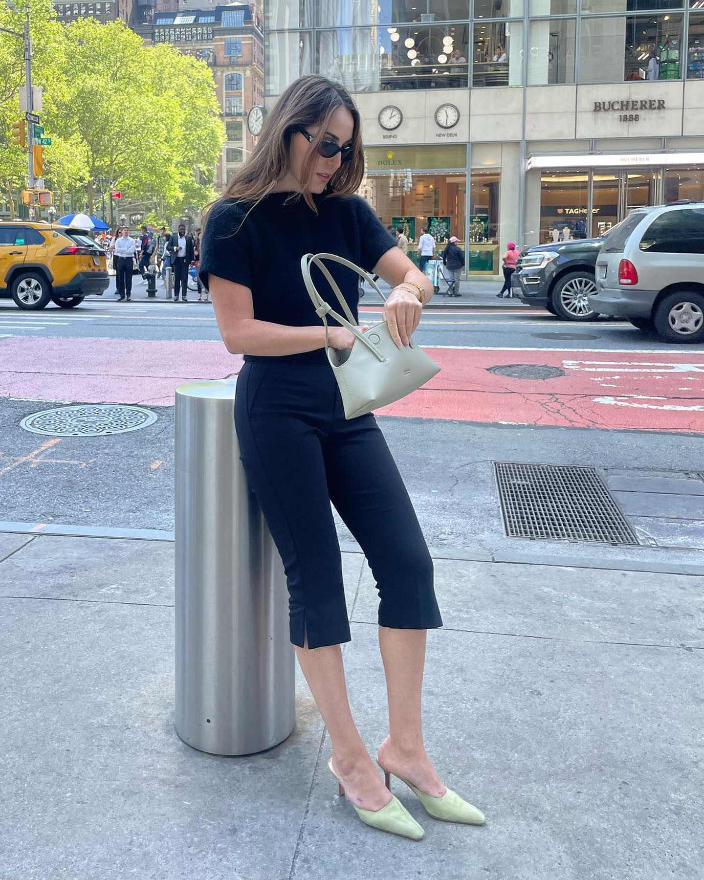 fashion editor Anna LaPlaca poses on the sidewalk in NYC wearing a short-sleeve black top, vented capri pants, a neutral shoulder bag, and light-green mule pumps