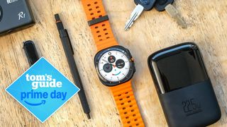 Samsung Galaxy Watch Ultra on an orange strap sitting on a wood table next to small accessories like keys, a pen and a power bank