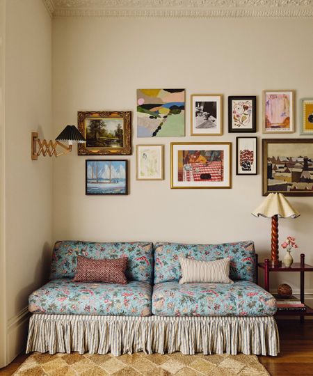A cozy corner featuring a blue floral-patterned sofa with a striped skirt, set against a cream wall adorned with an eclectic gallery of framed artwork and a vintage-style accordion wall lamp.