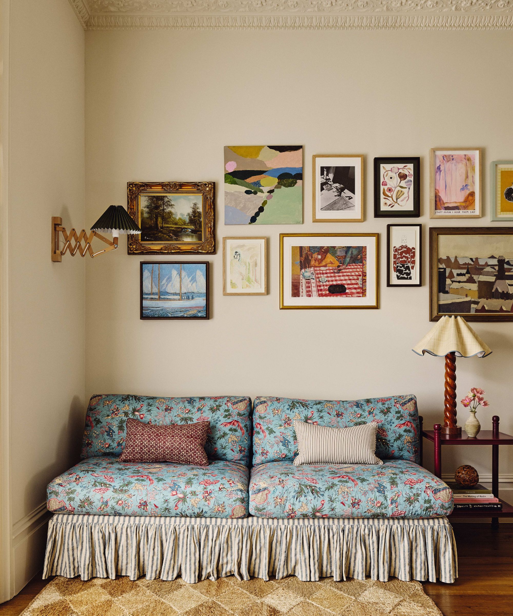 A cozy corner featuring a blue floral-patterned sofa with a striped skirt, set against a cream wall adorned with an eclectic gallery of framed artwork and a vintage-style accordion wall lamp.