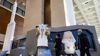 Two people walk by large models of China's moon rocket designs. A triple-core rocket, the Long March 10, stands the tallest.