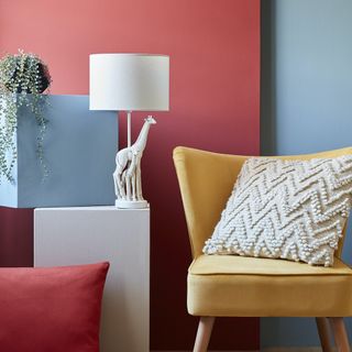 a yellow chair with a cream cushion in a corner where a red wall meets a blue wall, and white and blue artistic side tables next to it with a white giraffe lamp on top