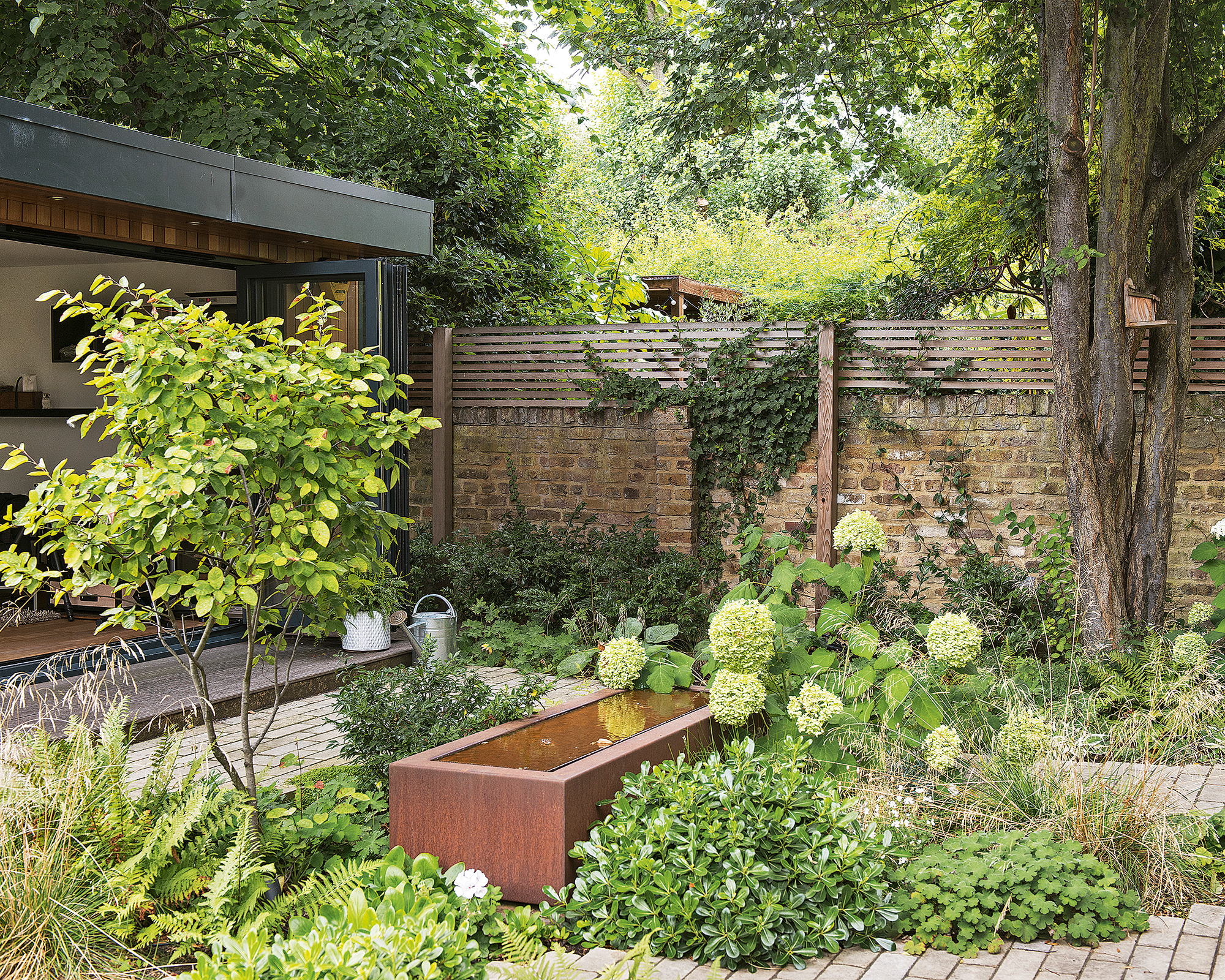 Small garden with brick wall, brick flooring, metal water container, flowers, bushes, trees